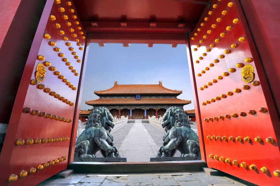 Grandes puertas rojas abiertas con pomos dorados enmarcan un palacio chino tradicional, custodiado por dos estatuas de leones de bronce.
