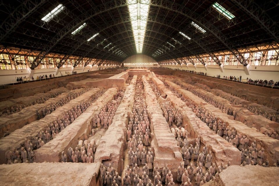 A wide, high-angle view of the Terracotta Army soldiers standing in formation in excavated pits inside a large hall.