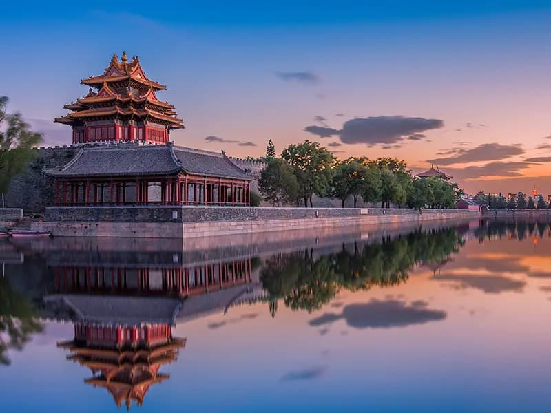 A traditional Chinese corner tower on a stone wall is reflected in the calm water of a moat at sunset.
