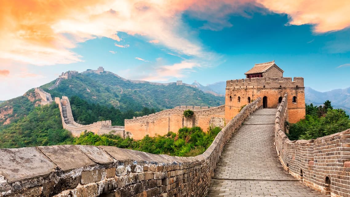A winding stone wall with watchtowers stretches across green mountains under a colorful sky at sunrise.