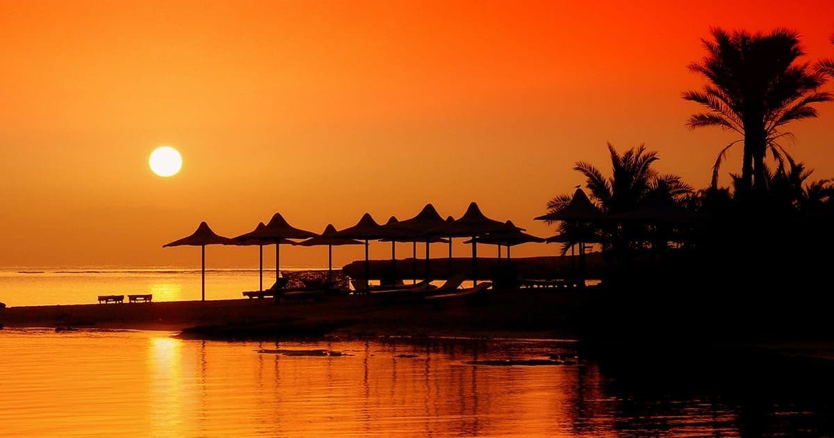 Silhouetten von Sonnenschirmen und Palmen an einem Ufer während eines orangefarbenen Sonnenuntergangs, wobei sich die Sonne auf dem ruhigen Wasser spiegelt.