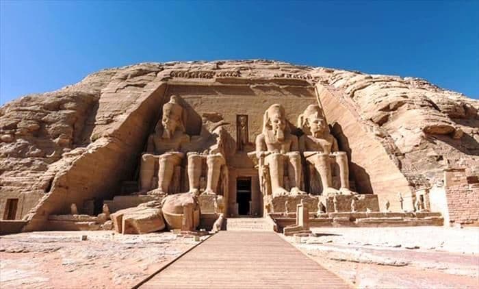 Una passerella di legno si avvicina all'ingresso di un grande tempio scavato nella roccia, affiancato da quattro colossali statue sedute.