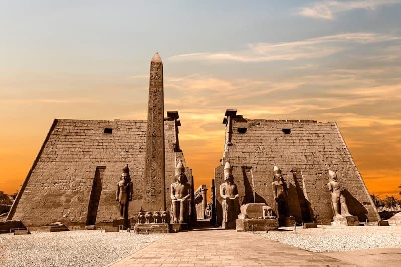 L'ingresso a un antico tempio egizio con un grande pilone, un obelisco e statue di faraoni sotto un cielo dorato al tramonto.