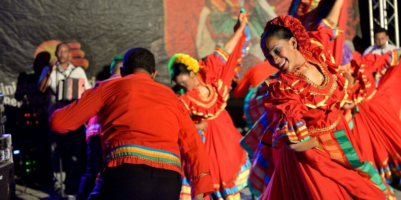 Des danseurs en costumes traditionnels rouges et colorés se produisent sur scène, avec une femme souriante au premier plan.