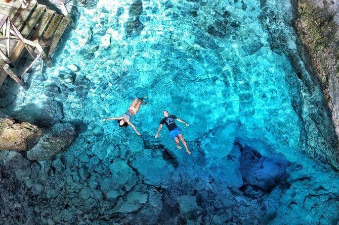 Vista aérea de un hombre y una mujer flotando de espaldas en aguas claras y turquesas sobre un fondo rocoso.
