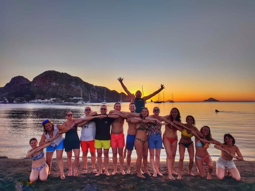 A WeRoad group trip poses with arms interlinked on a beach at sunset, with sailboats on the water behind them.