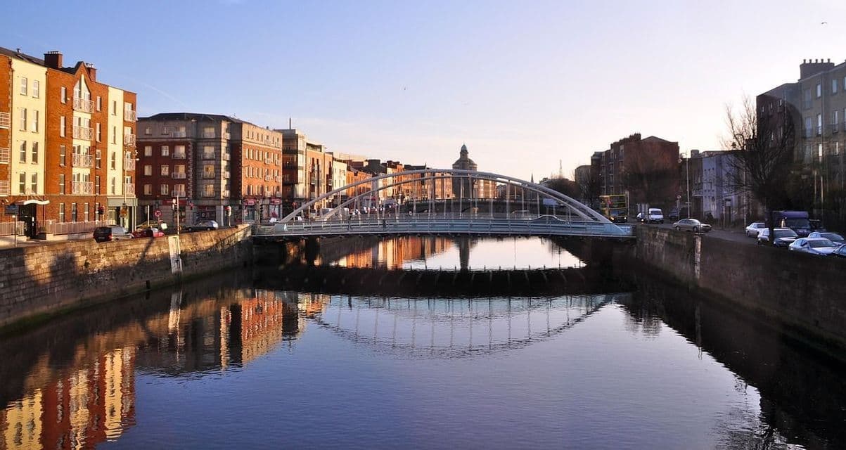 Un pont piétonnier en arc enjambe une rivière urbaine, avec son reflet et les bâtiments environnants visibles dans l'eau calme au coucher du soleil.