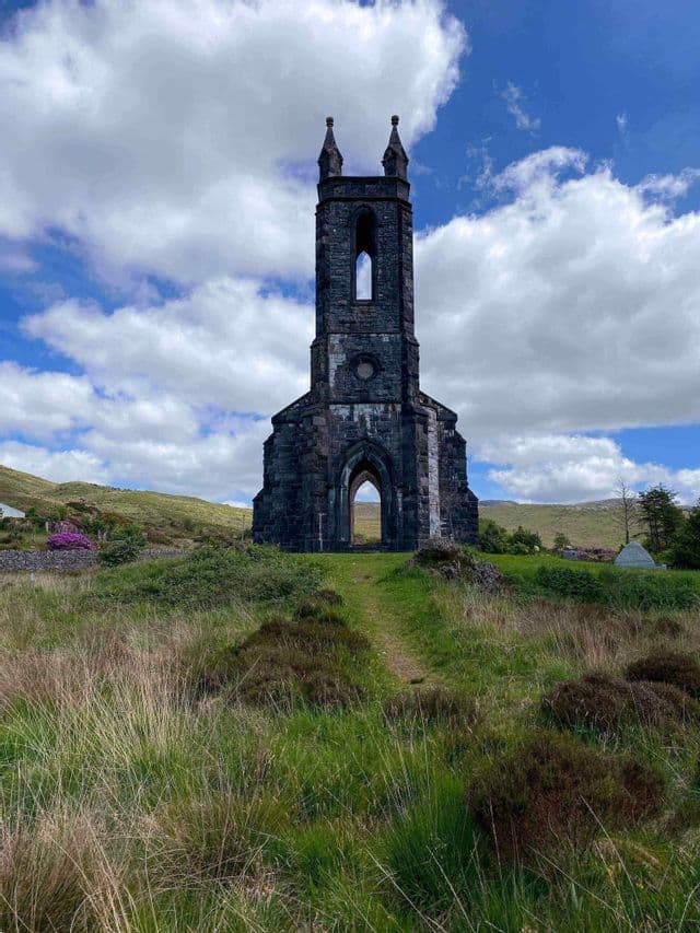 Una chiesa diroccata in pietra e alta si erge su una collina erbosa sotto un cielo blu con nuvole bianche.
