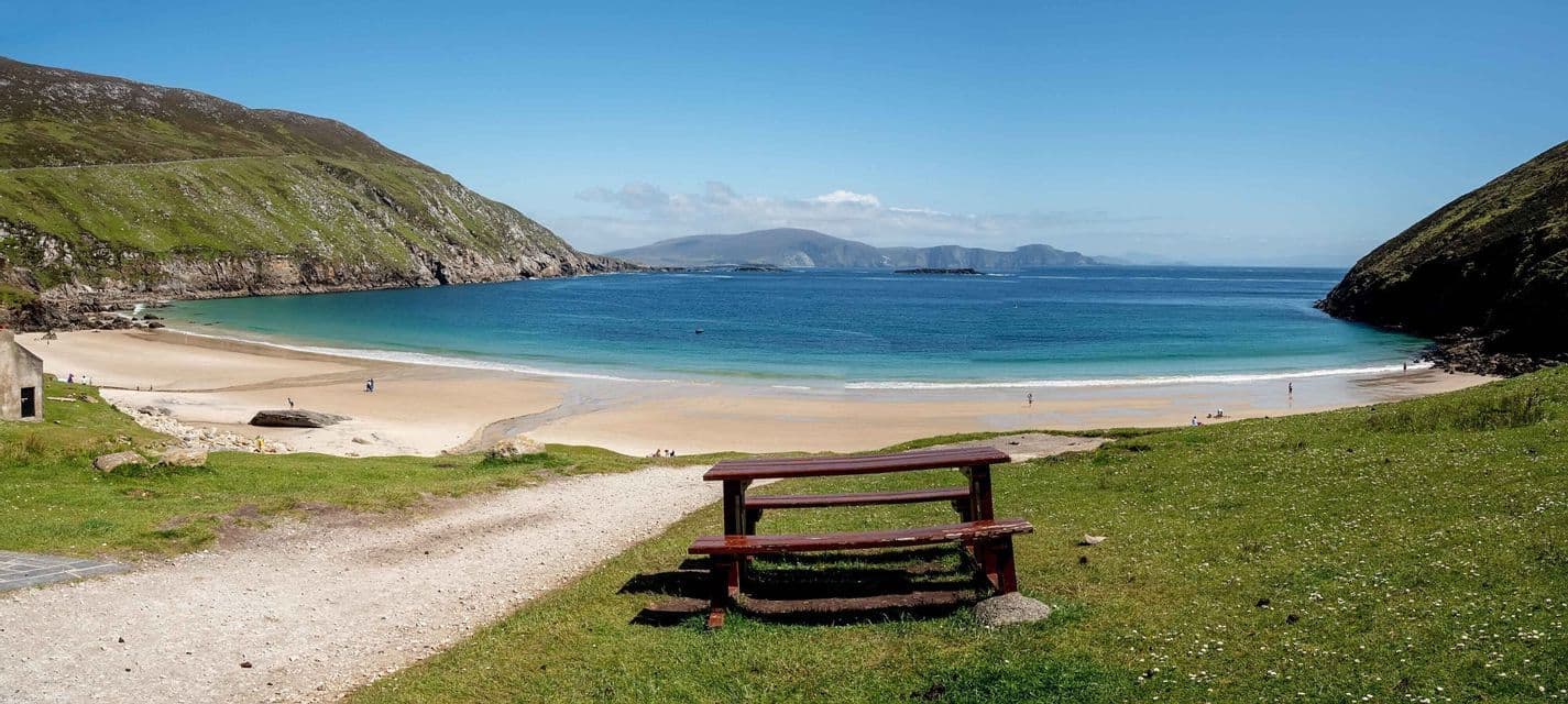 Un banc en bois sur une colline verdoyante domine une plage de sable isolée et une baie turquoise entre deux grandes falaises.