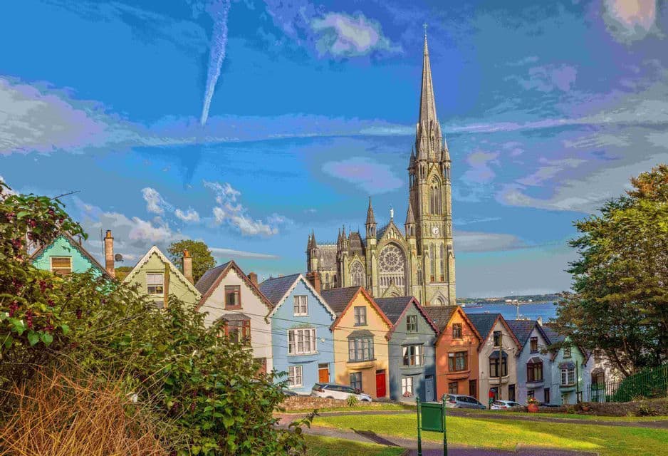 Une rangée de maisons colorées se trouve devant une grande cathédrale gothique en pierre, sous un ciel bleu parsemé de nuages.