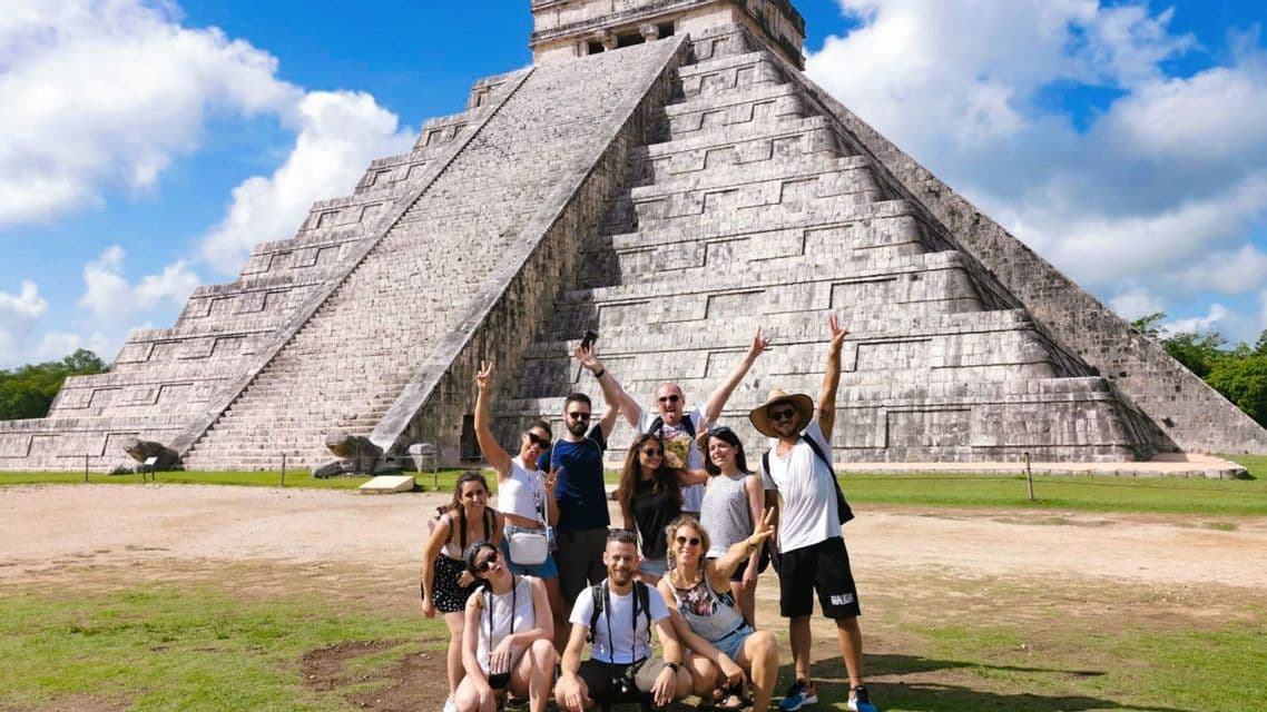 A WeRoad group trip posing for a photo on a grassy field in front of a large stone pyramid under a partly cloudy sky.