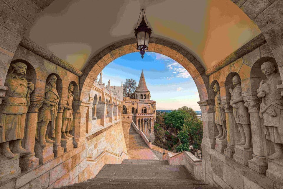 Una scala in pietra sotto un passaggio ad arco con statue di cavalieri, che si affaccia su una torretta di castello contro un cielo azzurro.