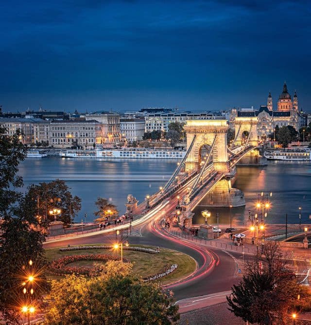 Eine beleuchtete Brücke über einen Fluss in der Dämmerung, mit Autoscheinwerfern, die in eine Stadt mit historischen Gebäuden führen.