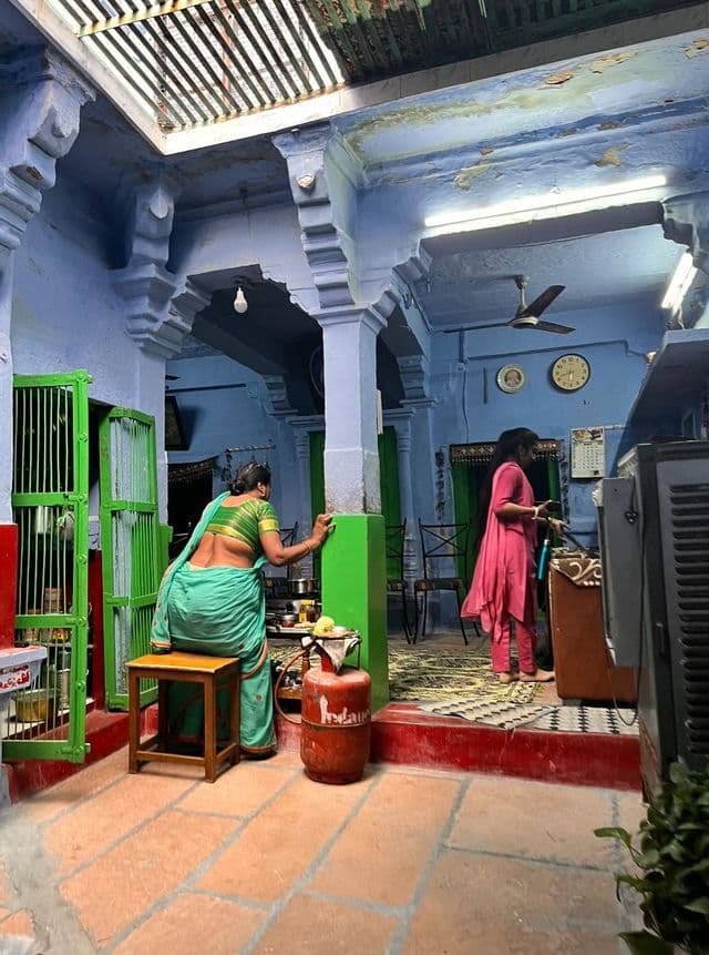 Una donna in sari verde cucina su un fornello a gas portatile all'interno di una casa con pareti blu ornate e pavimento in piastrelle di pietra.