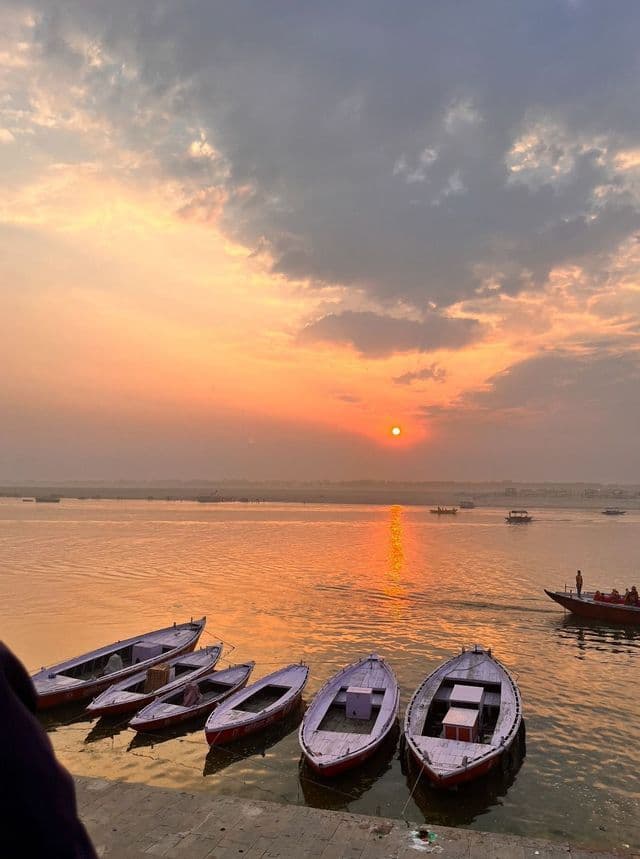 Mentre il sole tramonta, diverse barche viola sono ormeggiate su un fiume tranquillo, tingendo l'acqua e il cielo nuvoloso di un bagliore arancione.