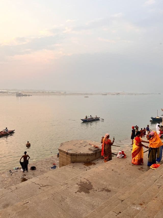 Al tramonto, persone con abiti colorati si ritrovano sui gradini in pietra di un ampio fiume, con alcuni che fanno il bagno e piccole imbarcazioni sull'acqua.