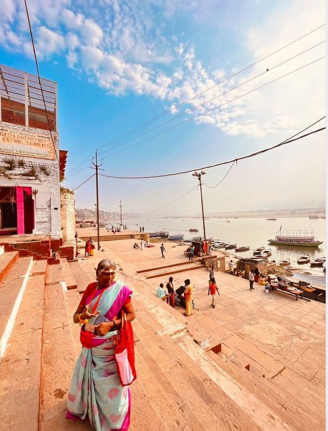 Una donna anziana con un sari colorato si trova su scalini in pietra che scendono verso un ampio fiume, con barche e persone lungo le sponde.