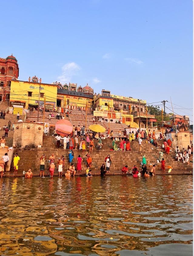 Un folto gruppo di persone che si bagnano in un fiume e stanno su gradini di pietra di fronte a edifici tradizionali e colorati sotto un cielo sereno.