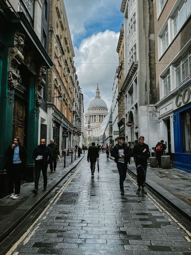 Des passants arpentent une rue pavée et mouillée d'une ville, le dôme d'une grande cathédrale étant visible au loin.