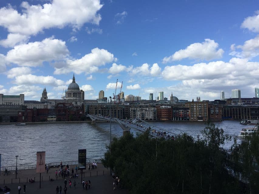 Vista di un paesaggio urbano con un fiume, un ponte pedonale affollato di persone e una grande cattedrale a cupola sullo sfondo sotto un cielo azzurro nuvoloso.