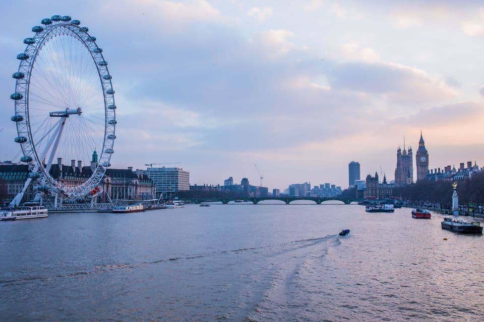 Un paysage urbain au crépuscule, avec une grande roue et une tour de l'horloge de part et d'autre d'un large fleuve sillonné de bateaux.