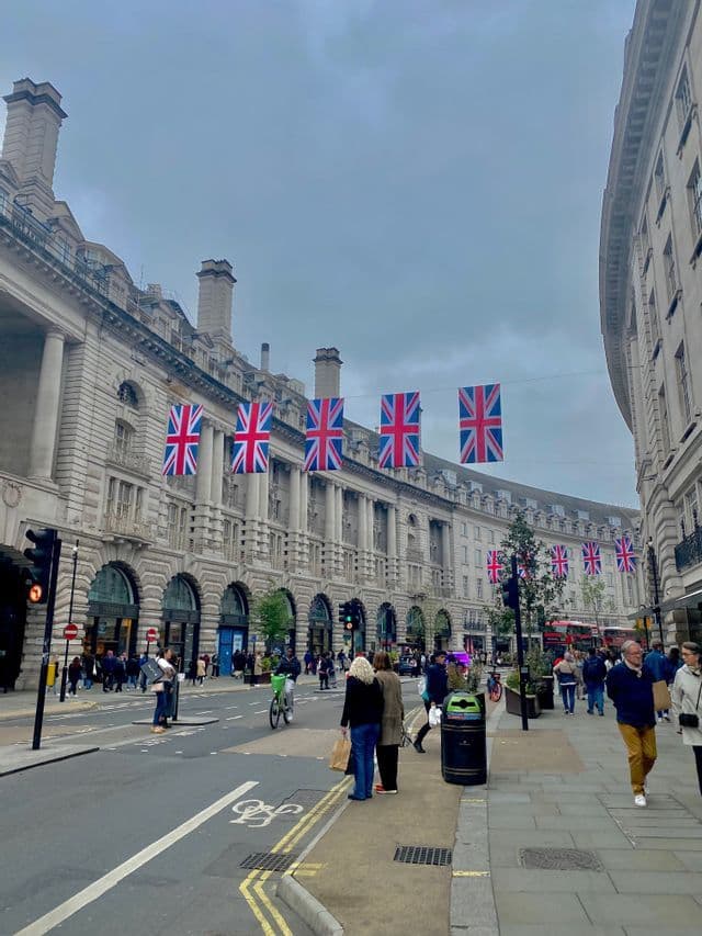Bandiere britanniche sono appese lungo una strada cittadina curva, fiancheggiata da architettura classica, mentre i pedoni camminano sul marciapiede.