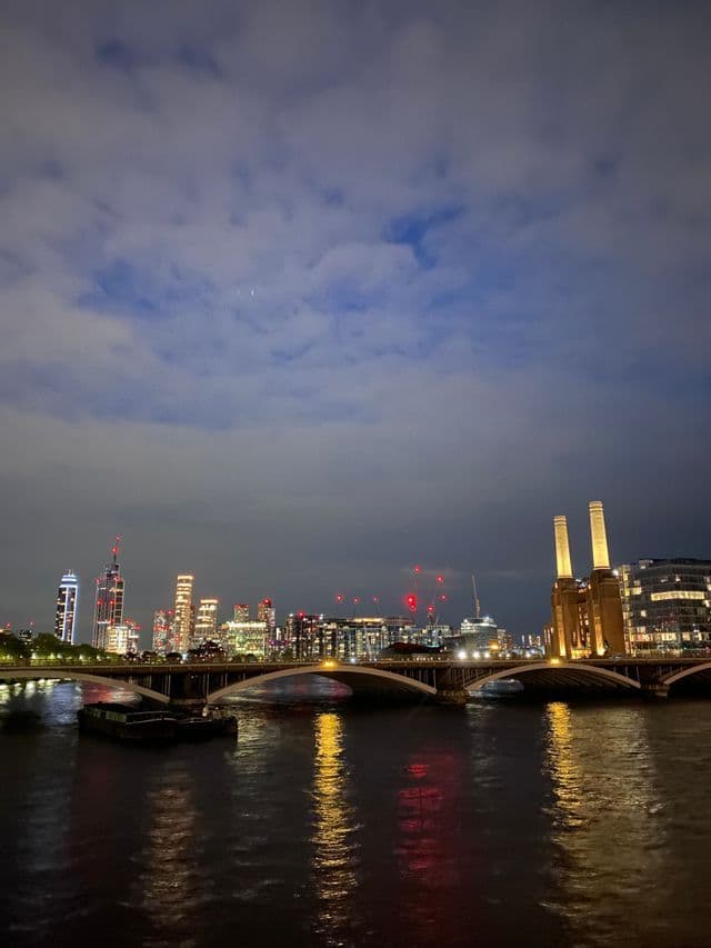 Une silhouette urbaine illuminée avec un pont et une centrale électrique se reflète dans une large rivière la nuit sous un ciel nuageux.