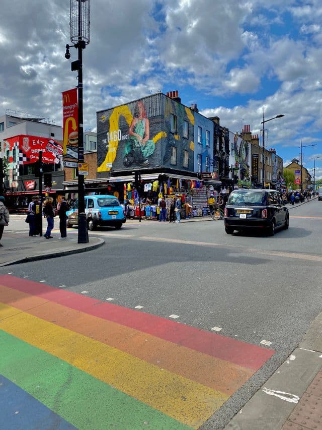 Un angolo di strada cittadina con un attraversamento pedonale arcobaleno, auto, pedoni ed edifici coperti da murales colorati sotto un cielo parzialmente nuvoloso.