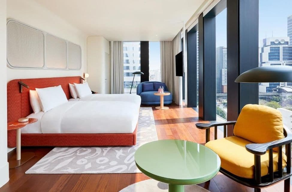 Ein sonnendurchflutetes, modernes Hotelzimmer mit zwei Betten, bunten Stühlen und raumhohen Fenstern mit Blick auf eine Stadtlandschaft.