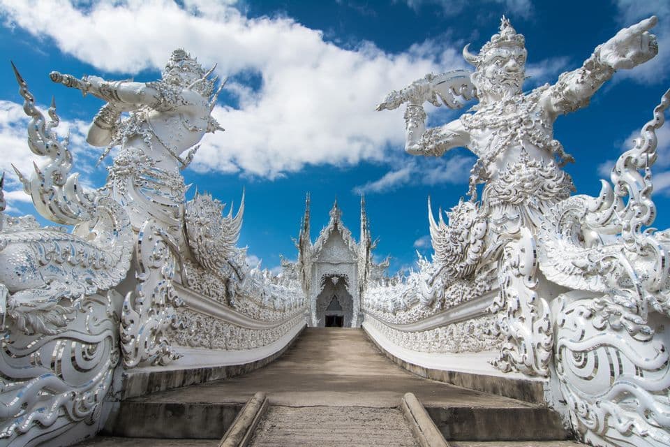 Vue en contre-plongée d'une allée menant à un temple blanc orné, encadré de grandes statues complexes sous un ciel bleu.