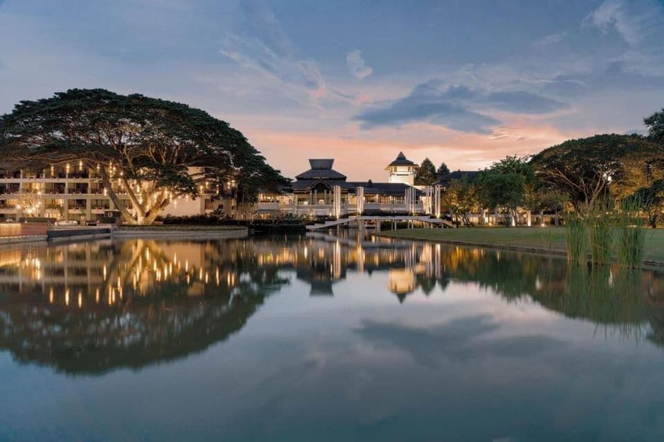 Un complexe hôtelier illuminé à l'architecture traditionnelle se reflète dans un lac calme au crépuscule, à côté d'un grand arbre décoré.
