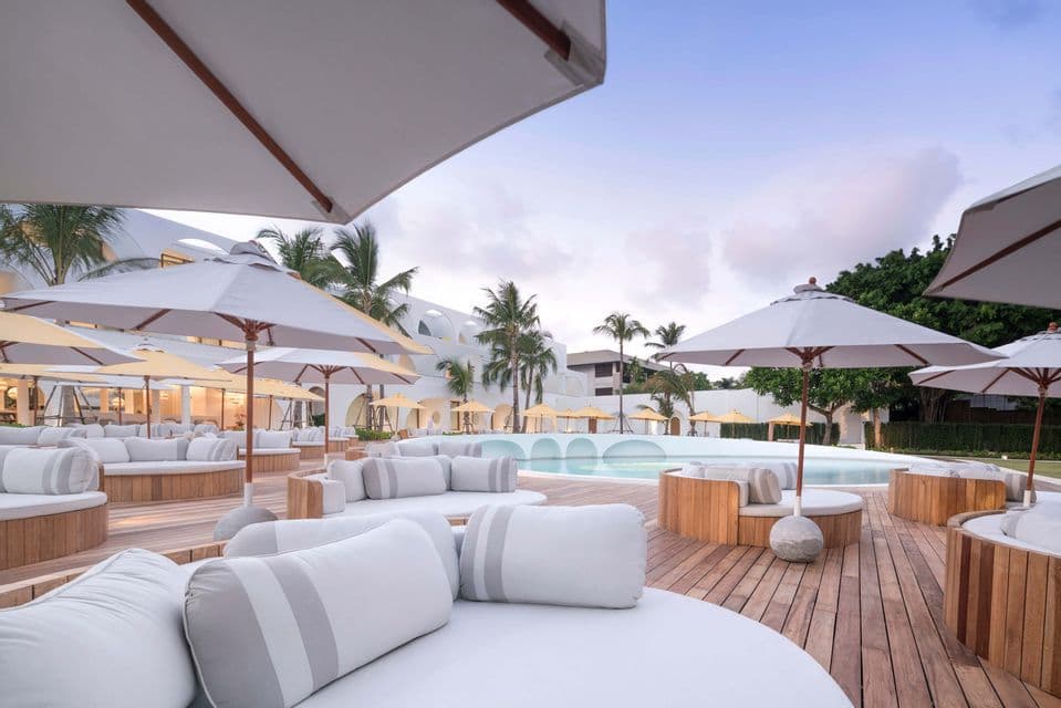 Una zona de salón al aire libre en un resort con tumbonas blancas y sombrillas en una terraza de madera junto a una piscina.
