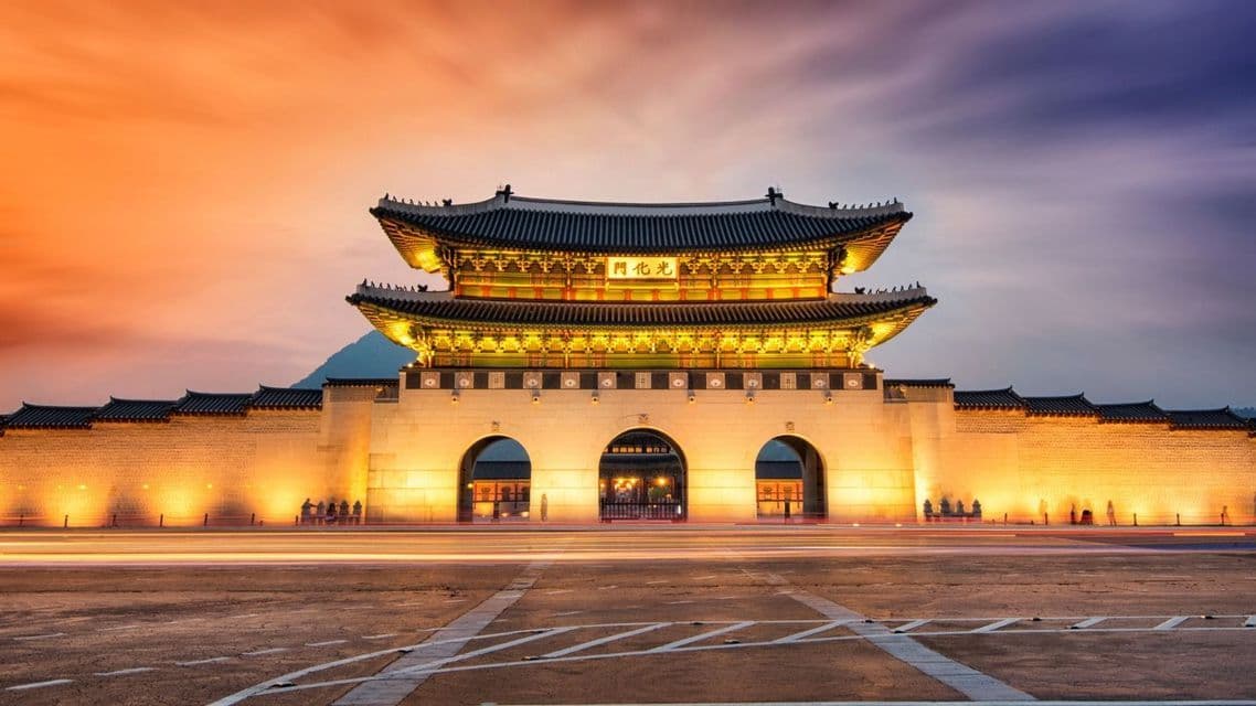 A large, illuminated traditional Asian gate at dusk, with long-exposure light trails from cars on the street in front.