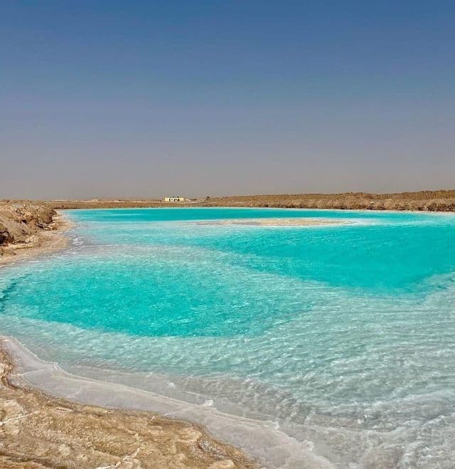 Ein leuchtend türkiser Salzsee mit einem weißen, kristallinen Ufer, umgeben von einer trockenen, braunen Landschaft unter klarem Himmel.