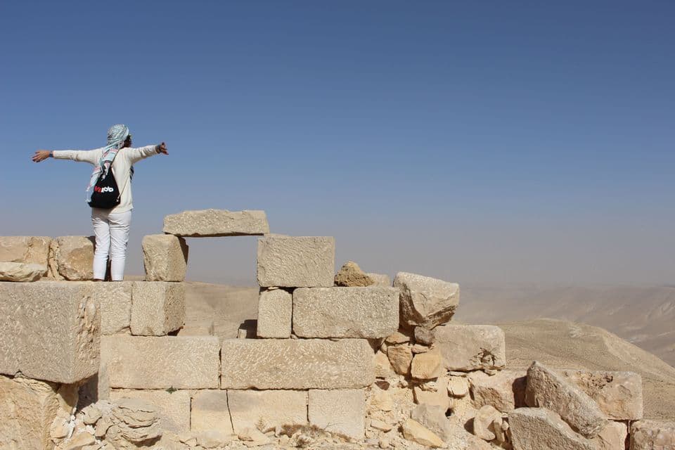 Ein Reisender auf einer WeRoad Gruppenreise steht mit ausgestreckten Armen auf steinernen Ruinen und überblickt eine Wüstenlandschaft.