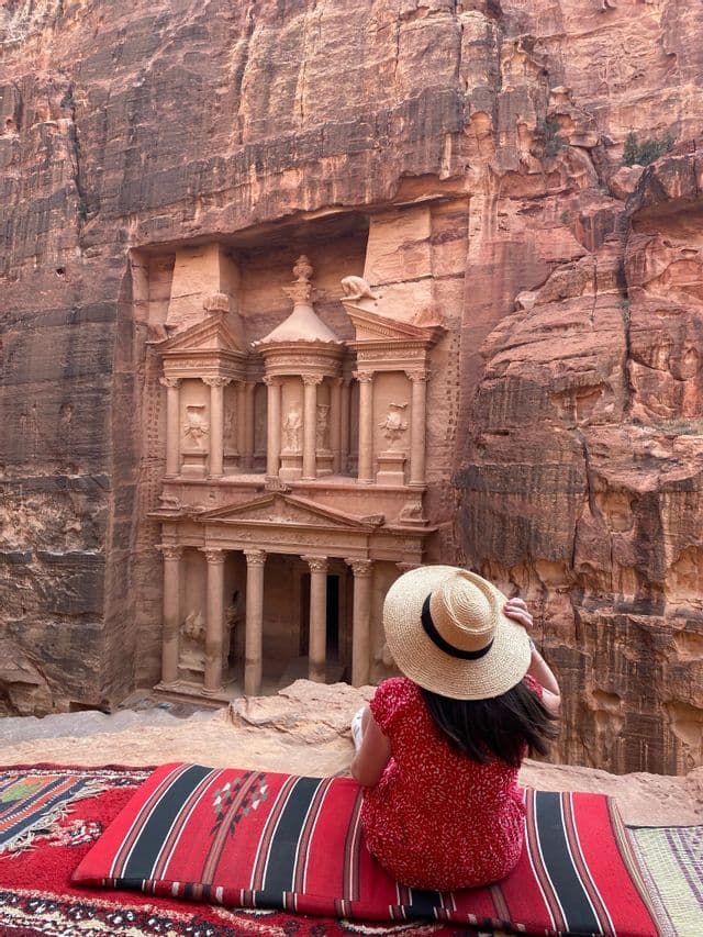Une femme en robe rouge et chapeau de paille est assise sur un tapis, admirant un grand temple sculpté dans une falaise de roche rouge.