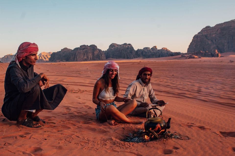 Una mujer de un viaje en grupo de WeRoad y dos hombres se sientan en dunas de arena roja, preparando té en una tetera sobre un pequeño fuego al atardecer.
