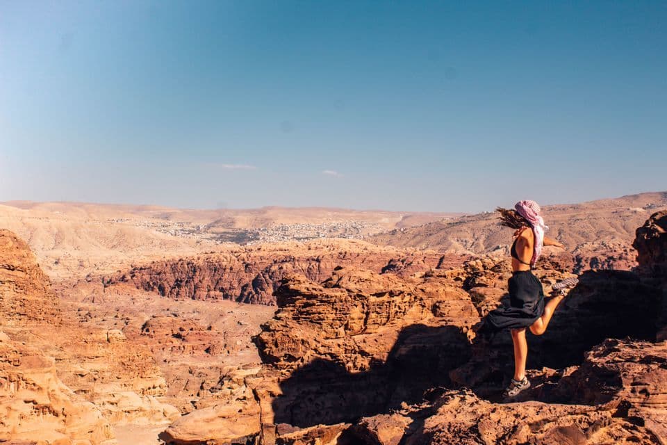 Una donna con un foulard posa su una gamba su uno sperone roccioso, affacciandosi su un vasto canyon desertico sotto un cielo azzurro e limpido.