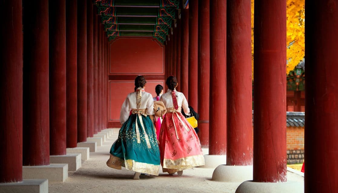 Tre donne viste di spalle, che indossano colorati abiti tradizionali hanbok, camminano lungo un corridoio con grandi pilastri rossi.