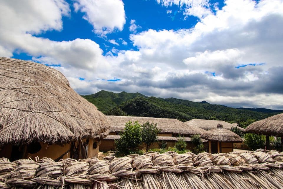 Un pueblo de cabañas tradicionales con techos de paja se encuentra al pie de montañas verdes bajo un cielo parcialmente nublado.