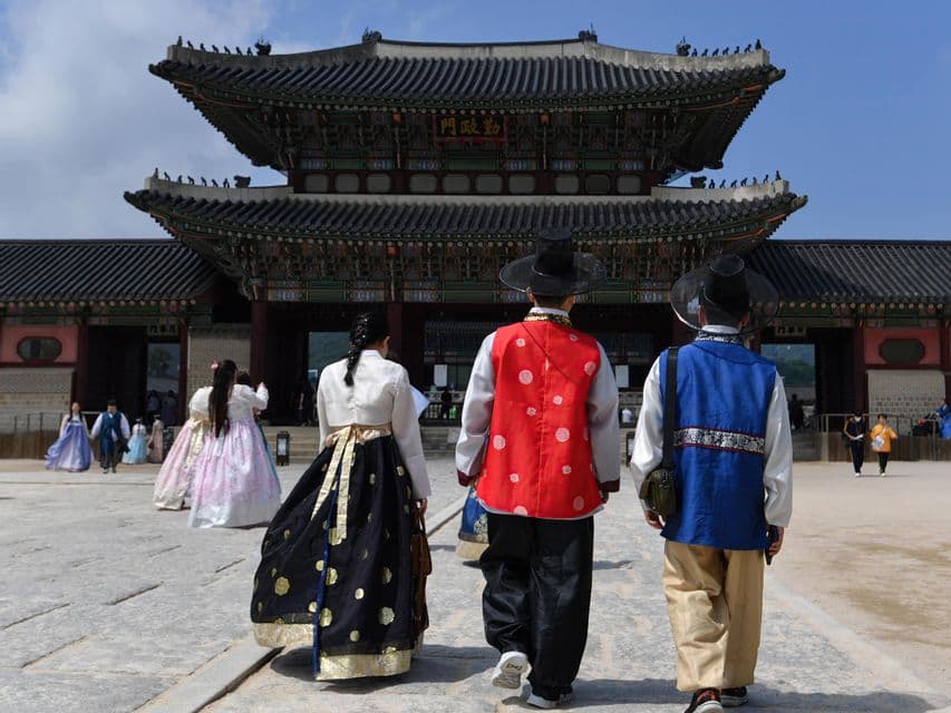 Drei Personen in farbenfroher, traditioneller koreanischer Kleidung sind von hinten zu sehen, wie sie unter klarem Himmel auf ein historisches Palasttor zugehen.