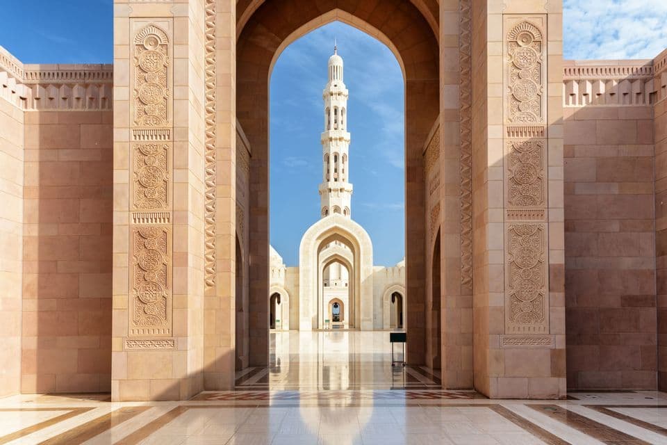 El minarete de una mezquita está enmarcado por un gran arco de piedra sobre un patio de mármol pulido bajo un cielo azul claro.