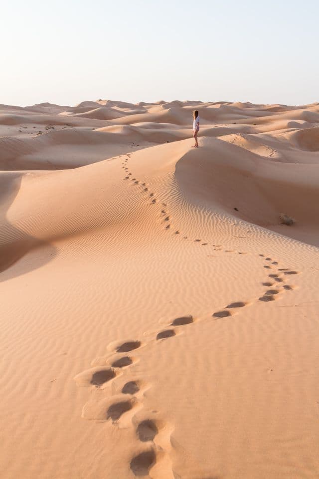 Une personne en chemise blanche se tient sur la crête d'une grande dune de sable, avec une traînée d'empreintes de pas menant jusqu'à elle.