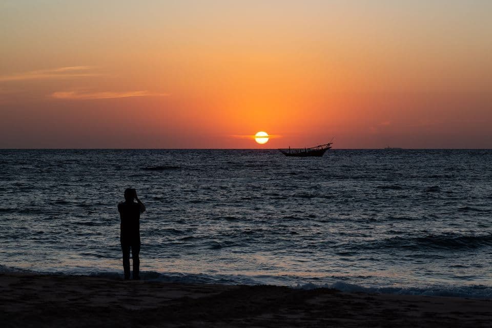 Una persona en silueta fotografía el atardecer naranja sobre el mar desde una playa, con un barco en el horizonte.