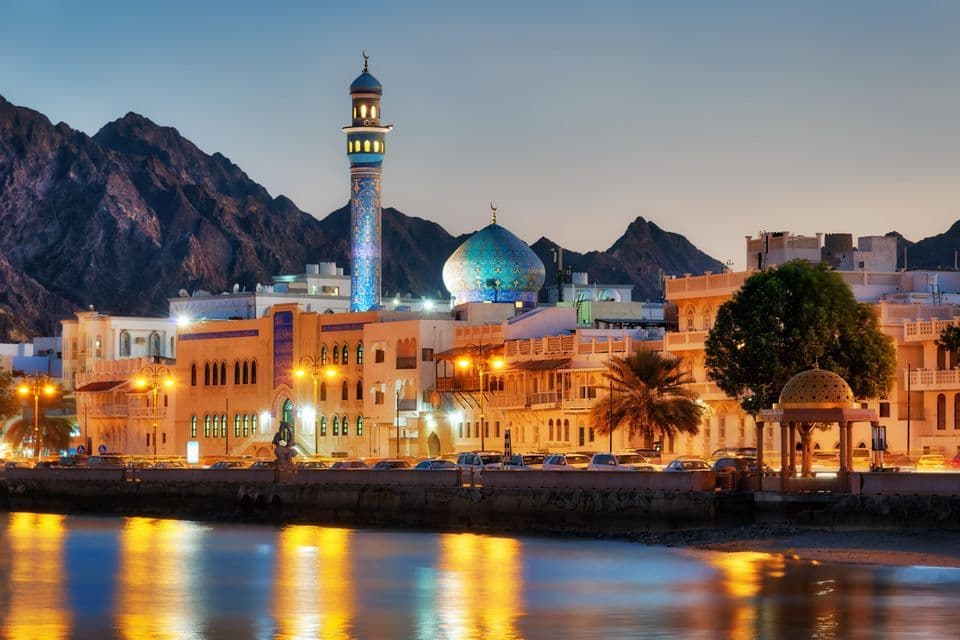 Une mosquée illuminée avec un dôme en mosaïque bleue et un minaret sur un front de mer, avec des montagnes derrière la ville au crépuscule.