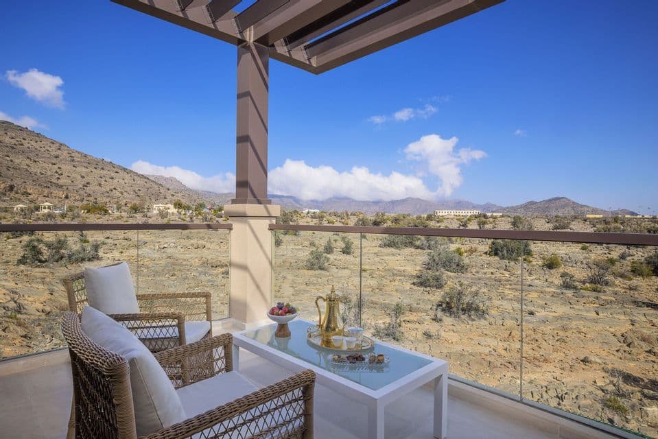 Un balcon avec deux chaises en osier et une table dressée avec un service à café, donnant sur un paysage montagneux et sec sous un ciel bleu.