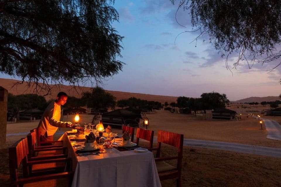 Un serveur allume une lanterne sur une table dressée pour un dîner en plein air dans un camp du désert au crépuscule.