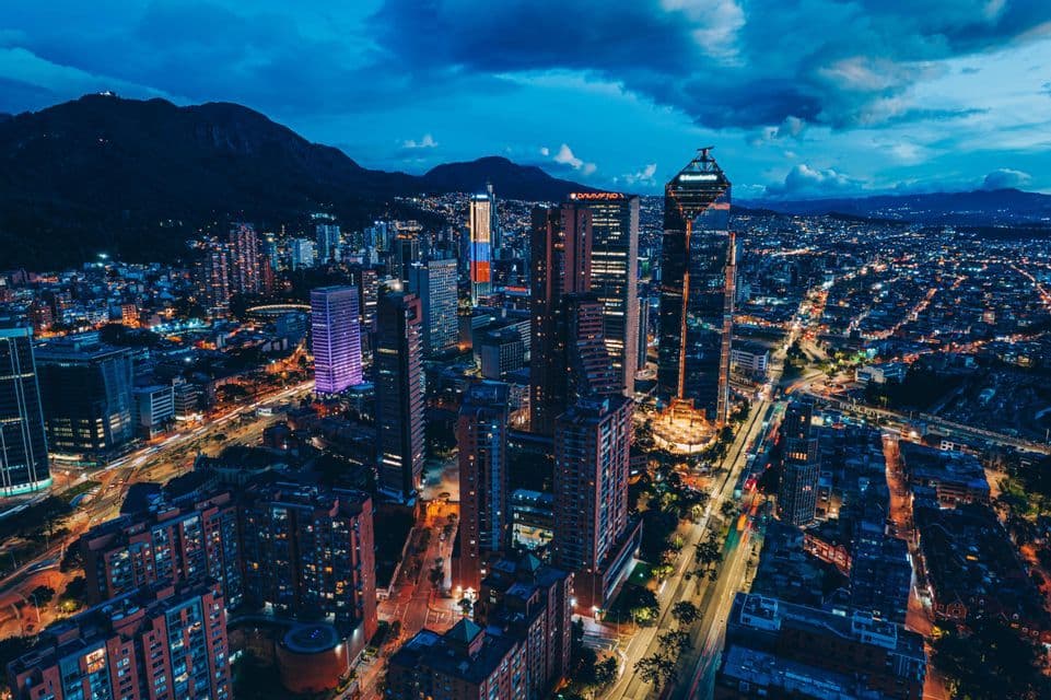 An aerial view of a sprawling cityscape illuminated by lights at dusk, with dark mountains silhouetted against a cloudy sky.