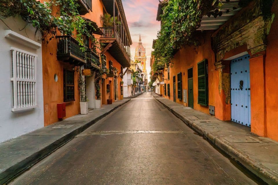 An empty street is flanked by colorful orange buildings with balconies and vines, with a church tower visible in the distance at sunset.