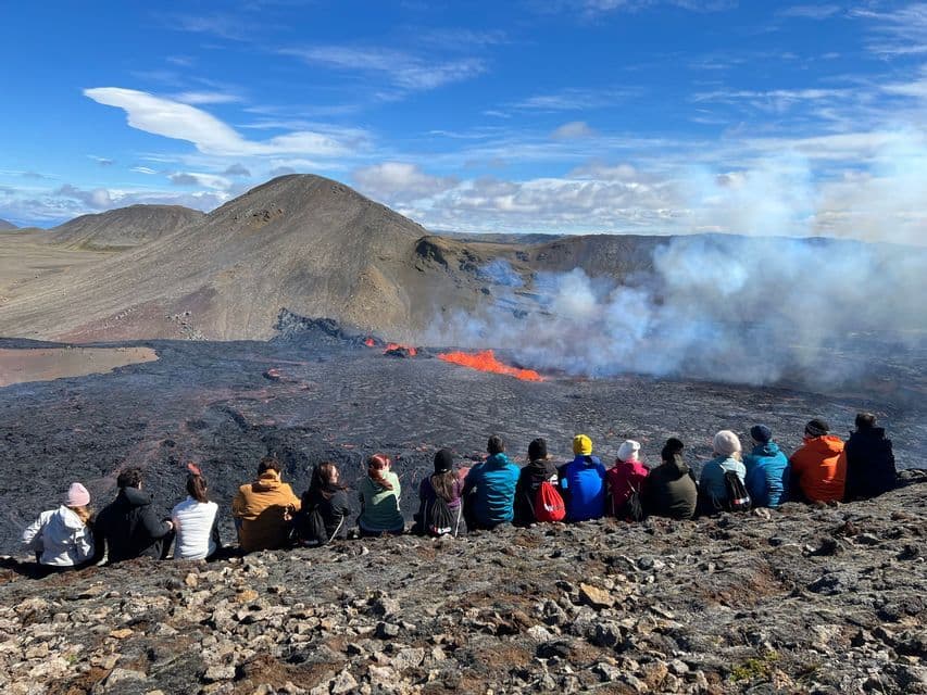 A WeRoad group trip seen from behind, sitting on a rocky hillside watching a volcanic eruption with flowing lava.